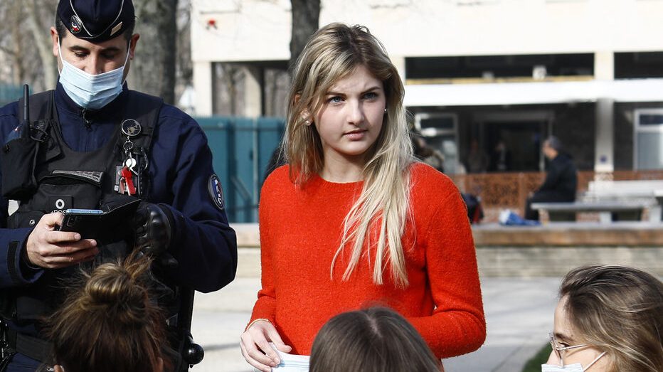 Thais d'escufon avec un pull rouge devant un bâtiment public, à sa droite un policier qui semble lui dresser une contravention Validisme / extrême droite / personnes handicapées